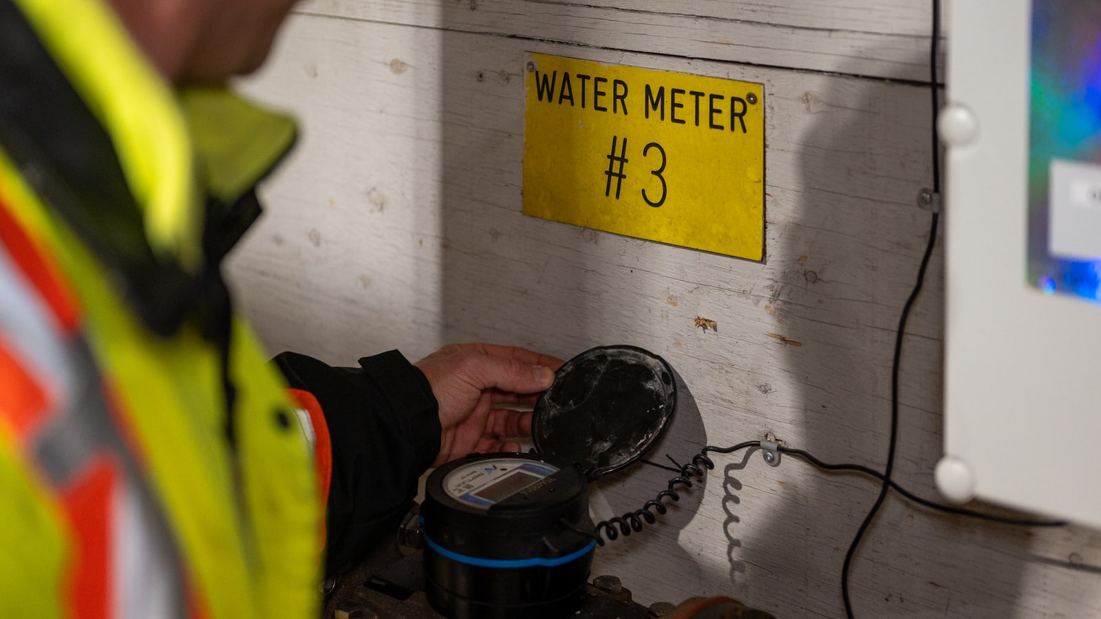 A worker in high-visibility clothing lifting the lid on a water meter, beside a Quantify enclosure
